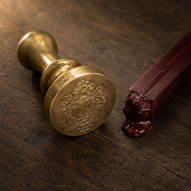 A heavy brass seal matrix lying beside a stick of dark red sealing wax on oak