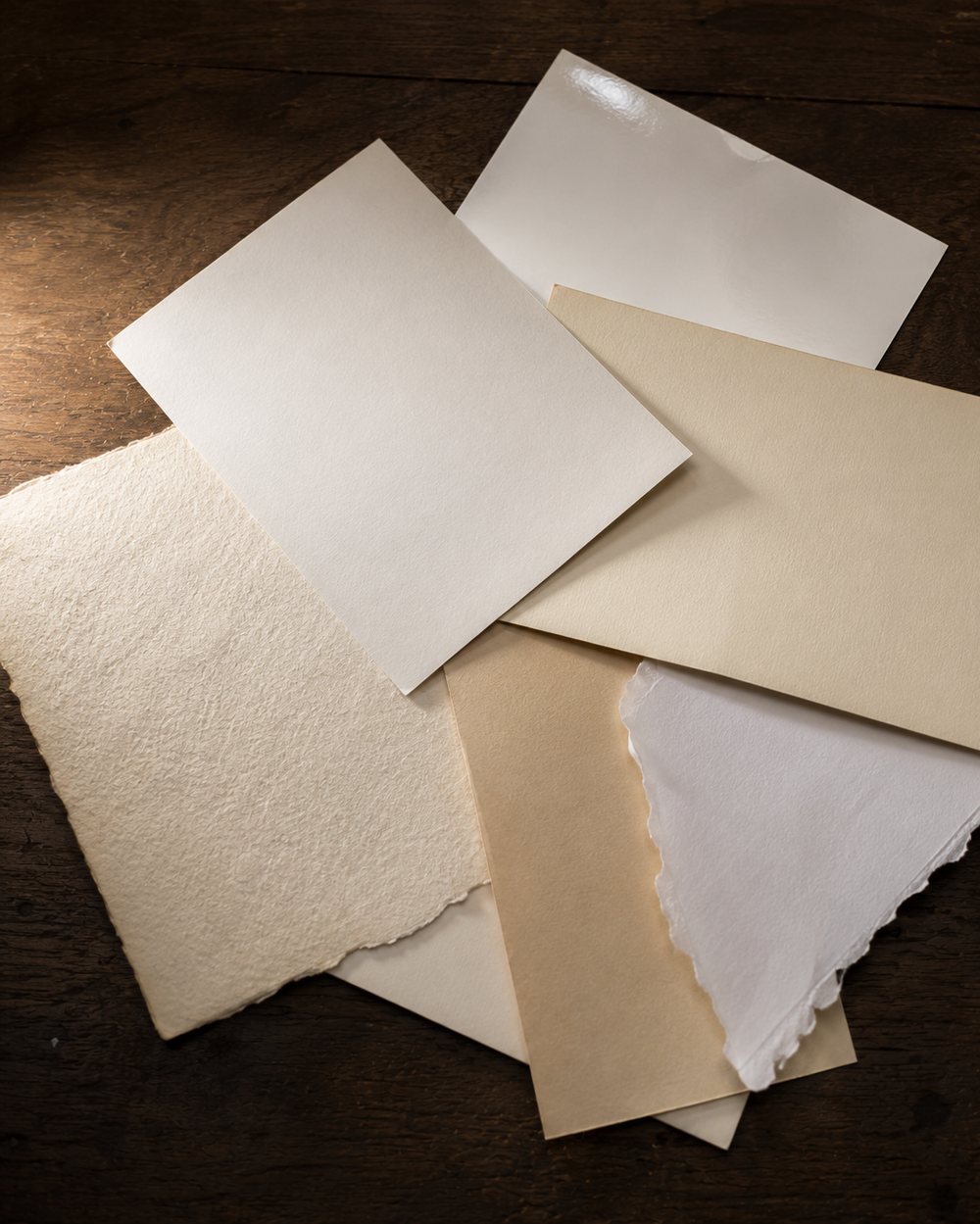 Blank paper scraps scattered on dark wood: different whites and creams, different thicknesses, one torn, one machine-cut, overlapping at clashing angles